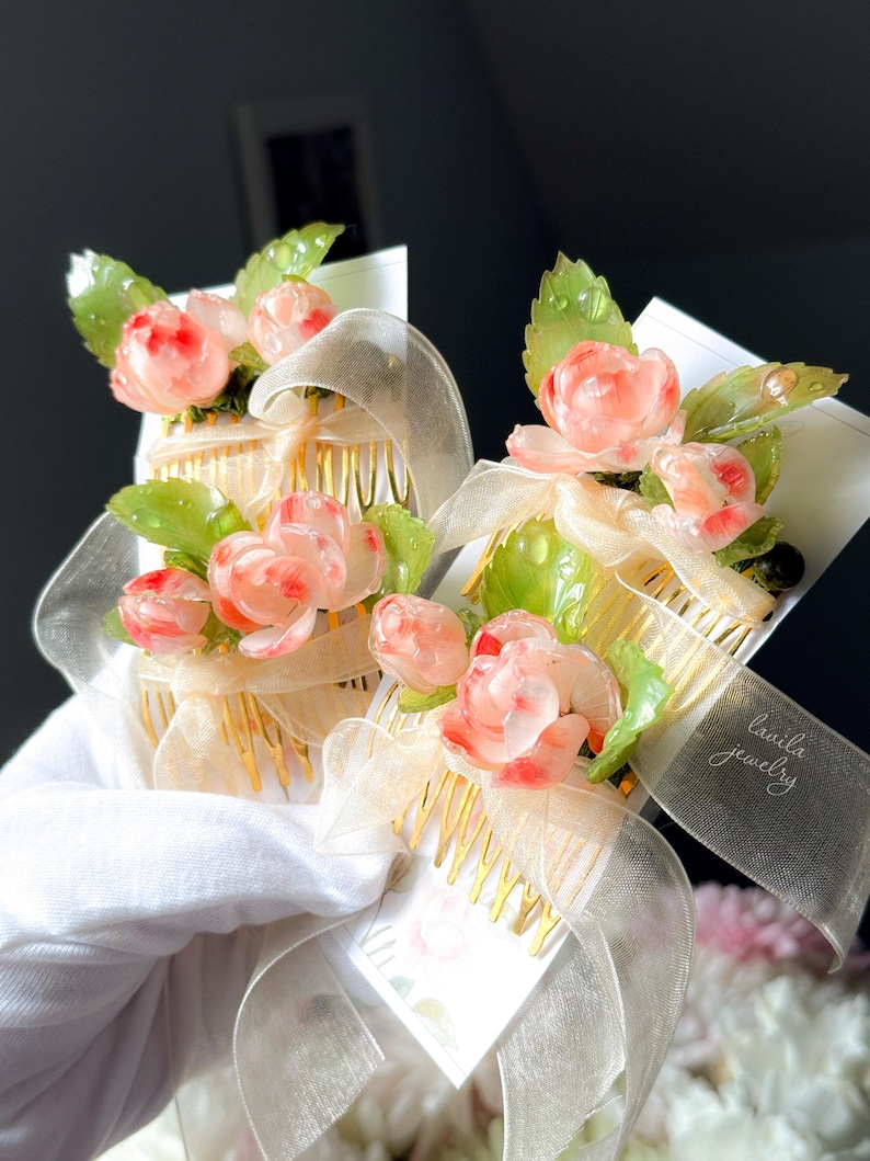 May include: Two floral hair combs with pink rose accents and green leaves. The combs have gold-colored teeth and are adorned with sheer ribbons. The text "lavila jewelry" is visible on one of the ribbons.