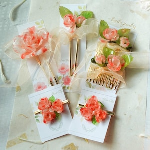 May include: Assortment of floral hair accessories, including hair clips, combs, and hair pins. Each piece features delicate, translucent peach-colored roses with green leaves. Some are adorned with gold-toned metal accents and ribbons. The accessories are displayed on cards.