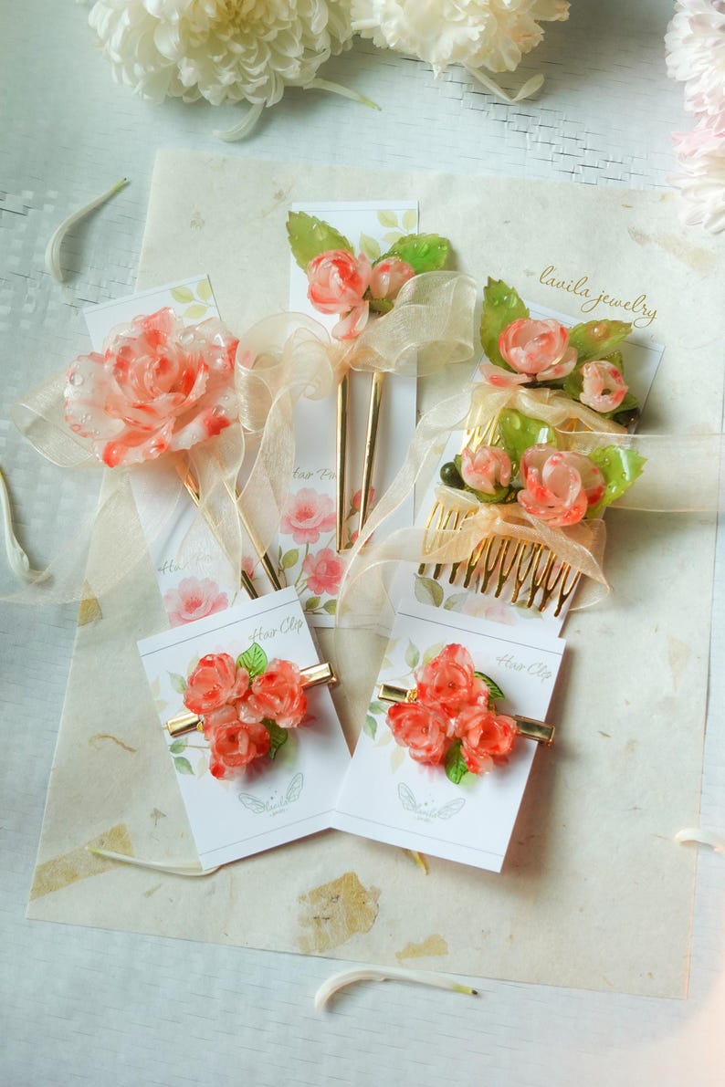 May include: Assortment of floral hair accessories, including hair clips, combs, and hair pins. Each piece features delicate, translucent peach-colored roses with green leaves. Some are adorned with gold-toned metal accents and ribbons. The accessories are displayed on cards.