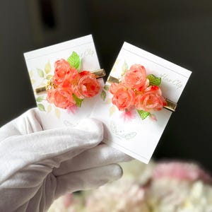 May include: Two floral hair clips featuring clusters of orange roses and green leaves. The flowers are attached to gold-colored metal barrettes. Each clip is displayed on a card with floral and butterfly designs.