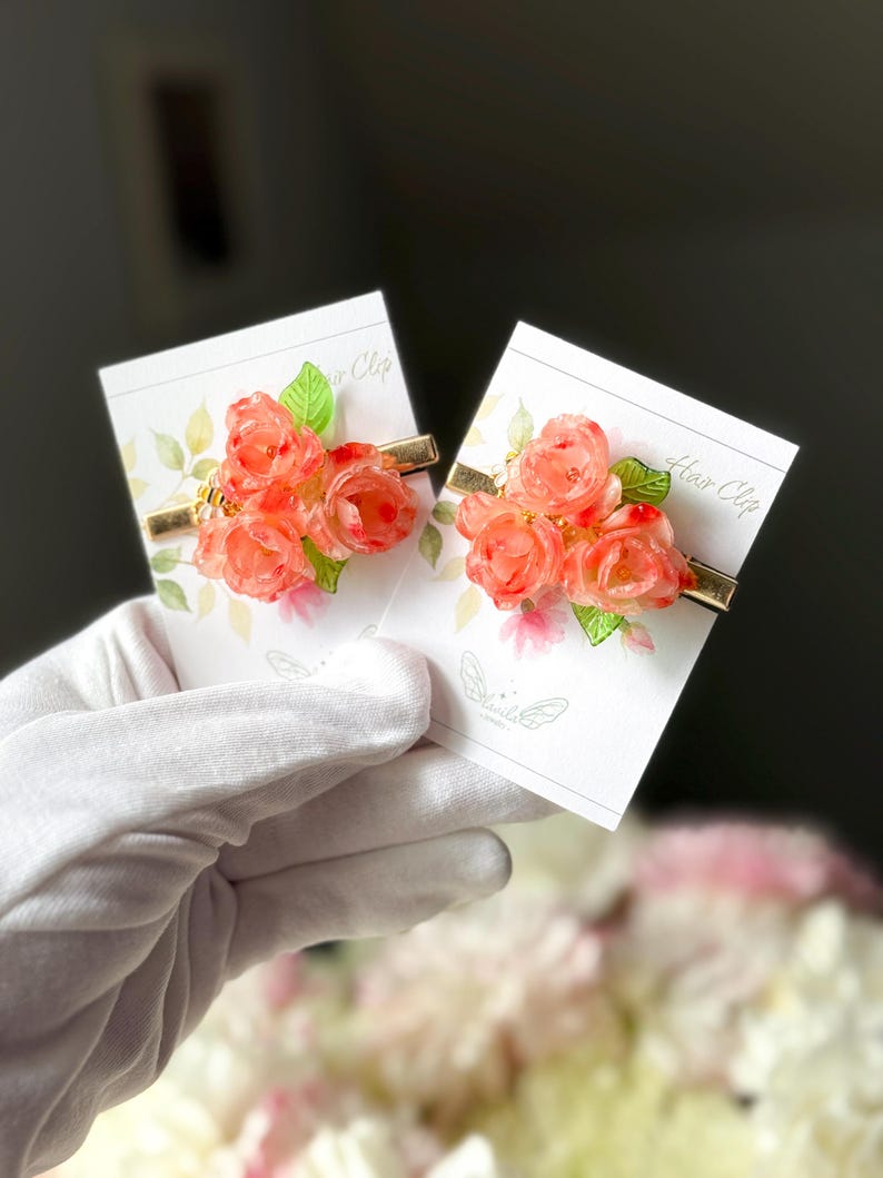 May include: Two floral hair clips featuring clusters of orange roses and green leaves. The flowers are attached to gold-colored metal barrettes. Each clip is displayed on a card with floral and butterfly designs.