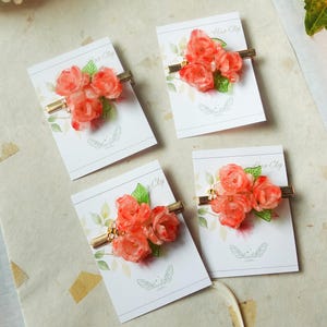 May include: Four hair clips with coral-colored rose clusters and green leaves on gold-tone metal bars. Each clip is displayed on a white card with the words "Hair Clip" and a butterfly logo.