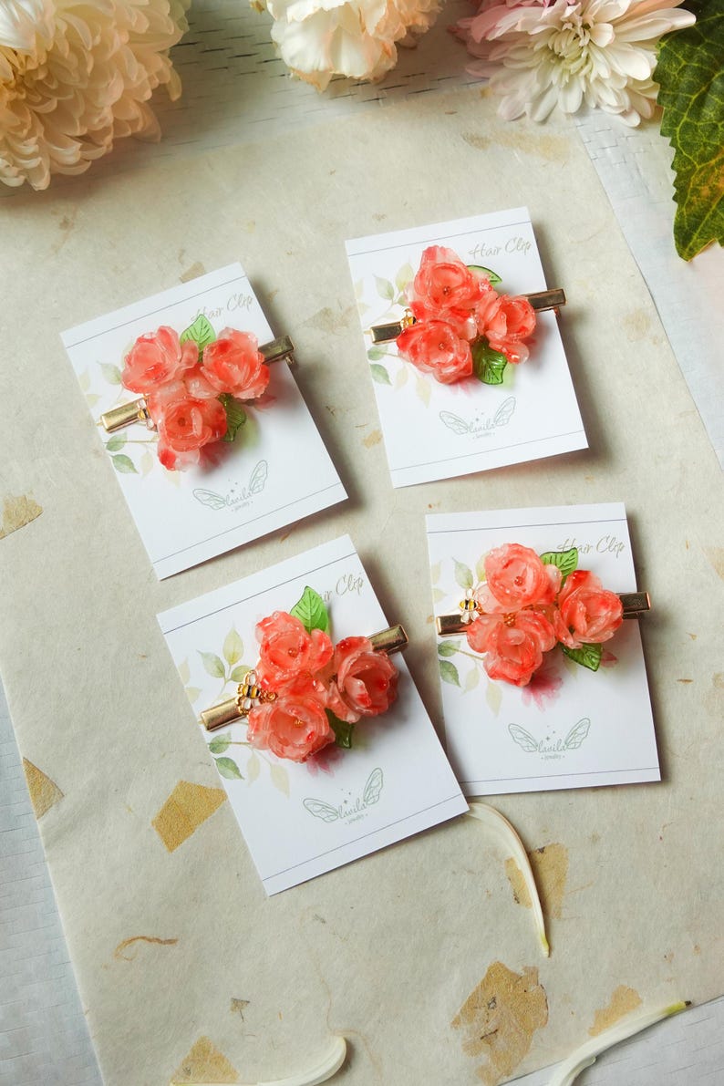 May include: Four hair clips with coral-colored rose clusters and green leaves on gold-tone metal bars. Each clip is displayed on a white card with the words "Hair Clip" and a butterfly logo.