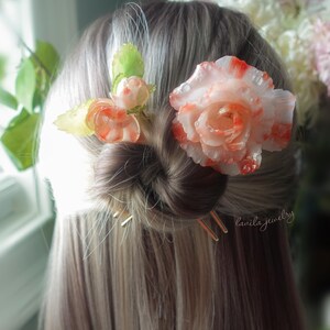 May include: A woman's hair styled in a bun, adorned with floral hair accessories. The hairpins are gold-colored. The flowers are white and orange, with green leaves. The text "lanila jewelry" is visible.