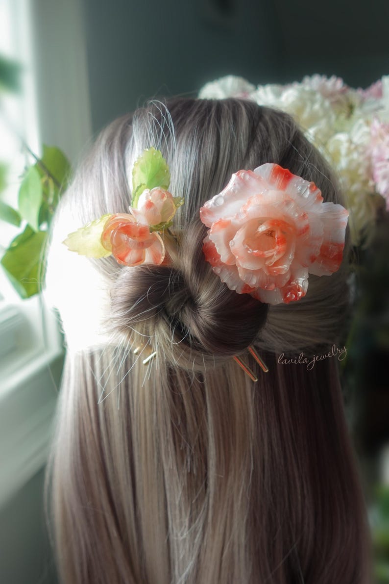 May include: A woman's hair styled in a bun, adorned with floral hair accessories. The hairpins are gold-colored. The flowers are white and orange, with green leaves. The text "lanila jewelry" is visible.