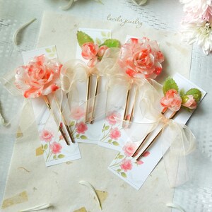 May include: Floral hair accessories featuring translucent orange and white rose designs with gold-toned hairpins. Each hairpin is adorned with a sheer ribbon and rests on a card with a rose illustration. The text "lavila jewelry" is visible.