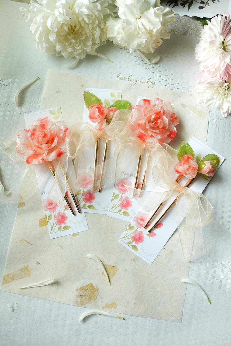 May include: Floral hair accessories featuring translucent orange and white rose designs with gold-toned hairpins. Each hairpin is adorned with a sheer ribbon and rests on a card with a rose illustration. The text "lavila jewelry" is visible.