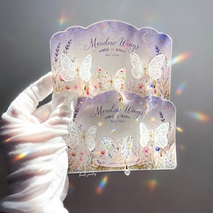 May include: Two display cards labelled Meadow Wings hair clips, each holding three translucent white butterfly hair clips with gold metal accents and small dangling iridescent crystal charms. The cards feature a floral and lavender illustration.