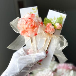 May include: A pair of white cards tied with a sheer ribbon, adorned with a large, translucent pink and red flower and smaller buds. The cards have a floral design and the text "lavila jewelry".