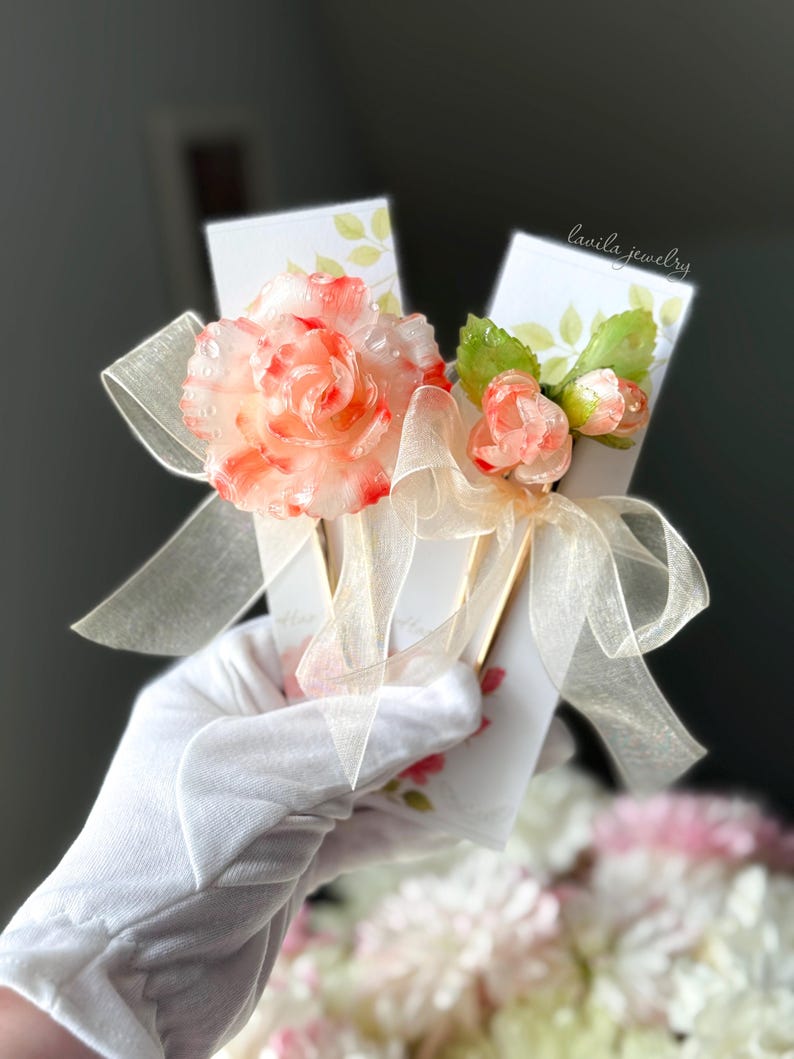 May include: A pair of white cards tied with a sheer ribbon, adorned with a large, translucent pink and red flower and smaller buds. The cards have a floral design and the text "lavila jewelry".