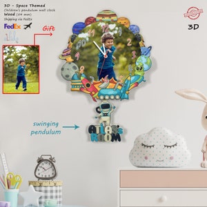 May include: A 3D wooden pendulum wall clock with a space theme. The clock features a colorful design with planets, rockets, and an astronaut. The clock face has a photo of a child in the center. The clock is 54 mm in diameter and has a swinging pendulum. The clock is labeled "ATLAS'S ROOM".