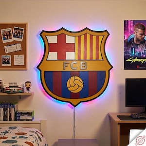 May include: Large, illuminated FC Barcelona team crest wall art. The wooden crest features the team's colours: red, blue, yellow, and white. The letters "FCB" are in the centre. The crest is outlined with a colourful LED light.