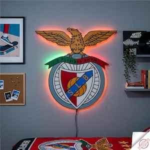 May include: Wooden Benfica emblem wall art with an eagle above a shield, illuminated by orange and green LED lights. The design includes the text "E Pluribus Unum" on a banner. The piece is connected to a power source via a black cable.