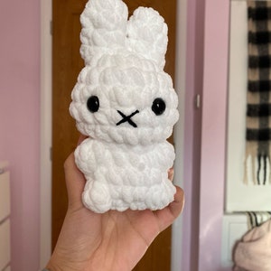 May include: A white, plush bunny toy with black eyes and a stitched black mouth. The bunny has large, upright ears and a textured, fluffy appearance. The toy is held in a hand, suggesting a soft, cuddly design.