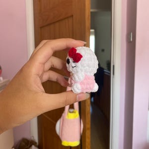 以下が含まれることがあります： ピンクのドレスと赤いリボンをつけた、小さな白い編みぐるみ。動物は黒い目で笑顔をしています。