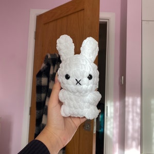 May include: A white, plush bunny toy with long ears and a stitched black mouth. The handmade stuffed animal has a textured, fluffy appearance. The bunny is held in a person's hand.