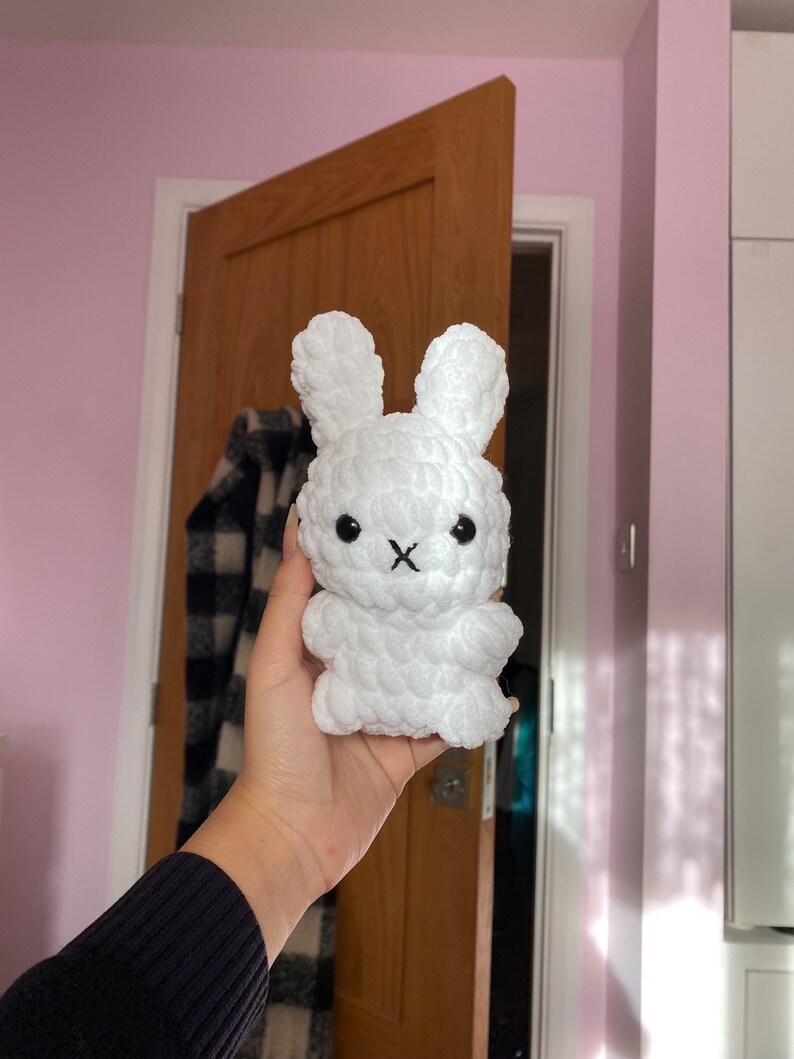 May include: A white, plush bunny toy with long ears and a stitched black mouth. The handmade stuffed animal has a textured, fluffy appearance. The bunny is held in a person's hand.