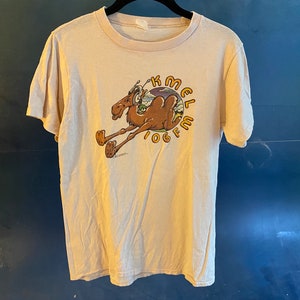 May include: A beige t-shirt with a cartoon camel wearing headphones and the text "K-MEL 106 FM" printed on the front.