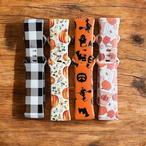 Pode incluir: Quatro pulseiras de relógio diferentes com vários padrões de outono. Uma pulseira é xadrez preto e branco, uma tem um design de abóbora e flores, outra tem um design de fantasma e morcego, e outra tem um design de latte de abóbora com especiarias.