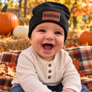 Peut inclure: Un bonnet en tricot noir avec un écusson rectangulaire en cuir marron portant le nom Hudson gravé en noir. Le bonnet est porté sur une tête, associé à un haut à manches longues en maille gaufrée crème avec trois boutons.