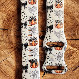 May include: A pair of white Apple Watch bands with a Halloween-themed print. The print features ghosts, pumpkins, bows, and spiderwebs in black and orange.