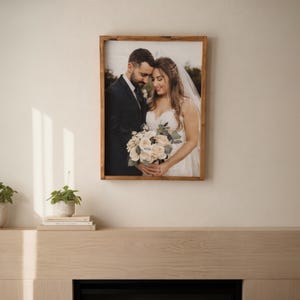 May include: A framed photograph of a wedding couple. The bride wears a white dress and veil, holding a bouquet of flowers. The groom is in a dark suit. The wooden frame hangs on a wall.