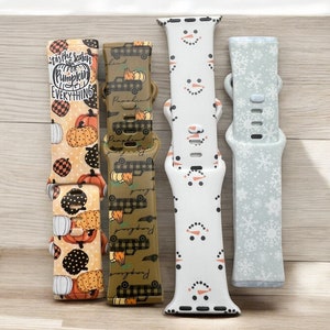 May include: Four different Apple Watch bands with various autumn and winter themed designs. The bands are in the colours of brown, green, white, and light blue. The designs include pumpkins, trucks, snowmen, and snowflakes.
