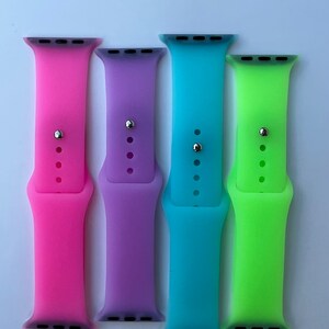 May include: Four silicone watch bands in pink, purple, blue, and green. The bands are designed for Apple Watches.