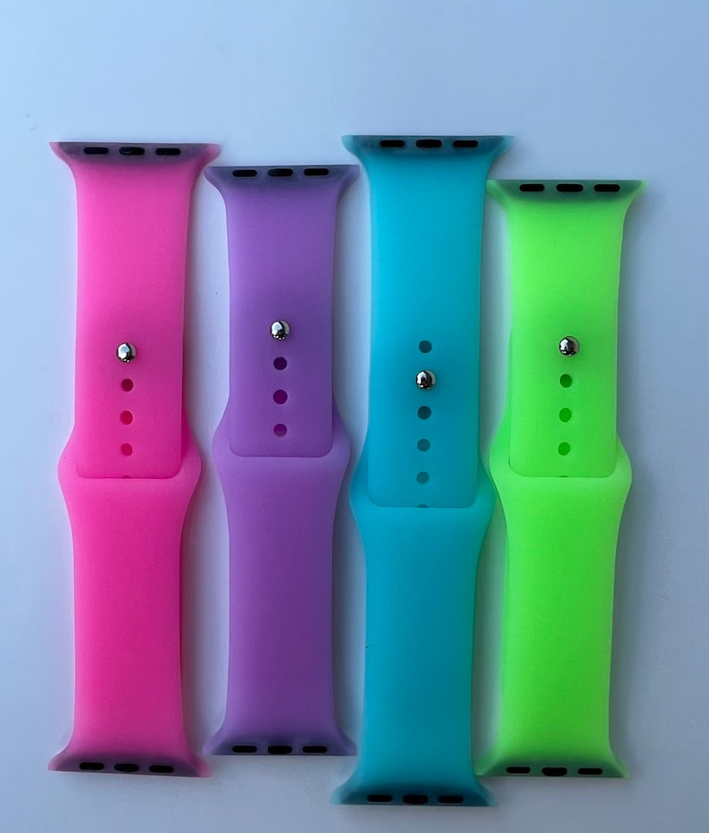 May include: Four silicone watch bands in pink, purple, blue, and green. The bands are designed for Apple Watches.
