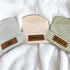 Puede incluir: Tres gorros de bebé a rayas con etiquetas de nombre de cuero personalizadas. Los gorros son de rayas azules, rosas y verdes. Los nombres en las etiquetas son Lily, Sophia y Banks.