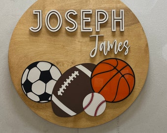May include: A round wooden sign with the name "JOSEPH JAMES" in white letters. Below the name are illustrations of a soccer ball, football, baseball, and basketball. The sign is light brown with a natural wood grain.