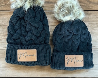 May include: Two black knit hats with faux fur pom poms. The larger hat has a brown leather patch that says "Mama" and the smaller hat has a brown leather patch that says "Mini".