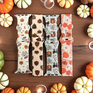 May include: Four Halloween watch straps with pumpkin, cow print, and leopard patterns. The straps are cream, tan, and orange, surrounded by miniature pumpkins on a wooden surface. The straps are for a watch.