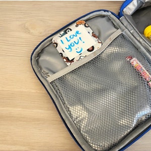 May include: A blue lunch bag with a mesh pocket and a note that says "I love you!" inside. The bag is open and there is a red and white candy bar inside.