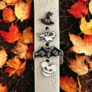 Könnte beinhalten: Ein weißes Uhrenarmband mit vier verschiedenen Halloween-Anhängern. Die Anhänger sind ein schwarzer Hexenhut, ein weißer Geist, eine schwarze Fledermaus und ein schwarz-weißer Mickey-Maus-Kopf.