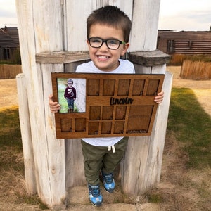 May include: A wooden photo frame with 12 slots for pictures. The frame has a small photo of a young boy in the top left corner. The frame is personalized with the name "Lincoln" and the words "First Birthday" and "Graduation".