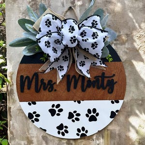 Puede incluir: Un letrero redondo de madera con un diseño blanco y marrón. El letrero presenta el texto "Mrs. Monty" en escritura negra, un patrón de huellas de patas y un lazo decorativo con huellas de patas.