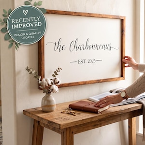 Puede incluir: Un letrero de pared rectangular con un marco de madera marrón oscuro y un fondo blanco. El texto dice "the Charbonneaus" en una fuente cursiva negra, seguido de "— EST. 2025 —" en una fuente negra sencilla.