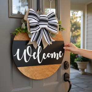 May include: A round wooden welcome sign with a black stripe and white script. The sign is adorned with a large black and white striped bow, burlap, and greenery. The sign is hanging on a gray door.