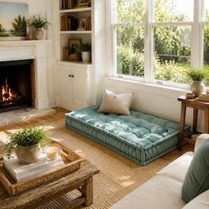 May include: A rectangular floor cushion in a muted teal color featuring a tufted surface with multiple fabric buttons. The cushion is placed on a woven rug and is topped with a single beige square throw pillow, providing a low-profile seating area.