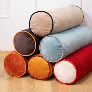 May include: A stack of six cylindrical bolster pillows in various colors including beige, dark brown, light blue, orange, yellow, and red. Each pillow features a contrasting circular end panel with piped edges, creating a soft, textured appearance.