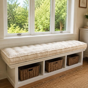 May include: A white wooden bench featuring a tufted cream-colored cushion on top and three open storage compartments underneath. Each compartment contains a rectangular woven brown wicker basket with a cutout handle for easy access and storage.