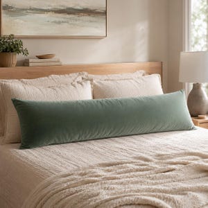May include: A long, rectangular bolster pillow in a solid sage green velvet fabric. The pillow is positioned horizontally across the front of a bed, resting against two cream-colored textured pillows and a matching cream-colored bedspread.