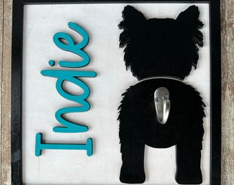 May include: A black wooden sign with a teal blue name "Indie" and a black silhouette of a dog's rear end with a silver hook attached.