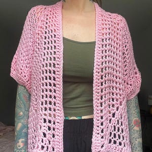 Può includere: Un cardigan all'uncinetto rosa chiaro a trama aperta con maniche corte. Il cardigan è realizzato in un filato morbido e strutturato e ha una vestibilità rilassata. L'indumento è indossato sopra una canotta verde e pantaloni neri.