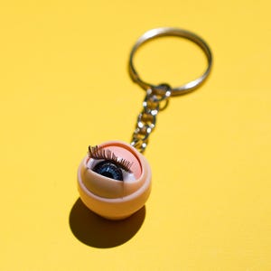 May include: A novelty keychain featuring a miniature eye with long eyelashes. The eye is set in a light peach-colored sphere, attached to a silver chain and keyring. The background is a solid yellow.