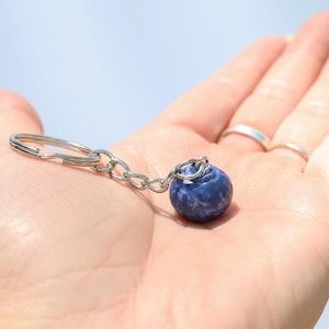 May include: A small, round, dark blue stone keychain with a silver-colored chain and key ring. The stone has a textured surface and is held in an open hand against a light blue background.