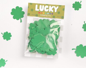 Puede incluir: Una bolsa de confeti verde con forma de trébol con la palabra "LUCKY" impresa en la parte superior. El confeti está esparcido sobre un fondo blanco con formas de trébol verdes adicionales.