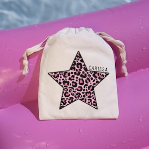 May include: A small off-white drawstring pouch featuring a large five-pointed star graphic with a pink and black leopard print pattern. The name CARISSA is printed in black capital letters on the right side of the star graphic.