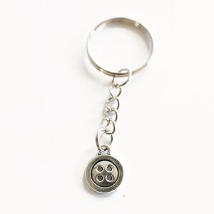 May include: Silver-toned keychain with a button charm. The button charm has four small circles in the center. The keychain is made of metal and is on a white background.